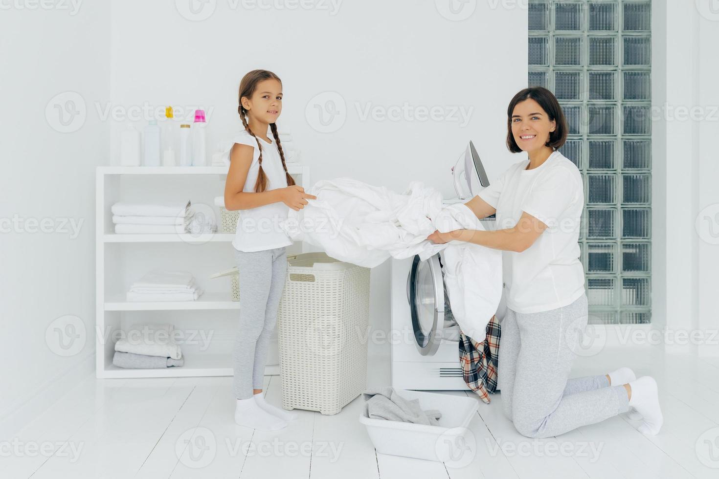 heureuse femme au foyer fait la lessive avec une petite aide adorable. la mère et la fille lavent les vêtements dans la buanderie, chargent le linge dans la laveuse. la femme se tient à genoux près de la machine à laver. notion de ménage photo