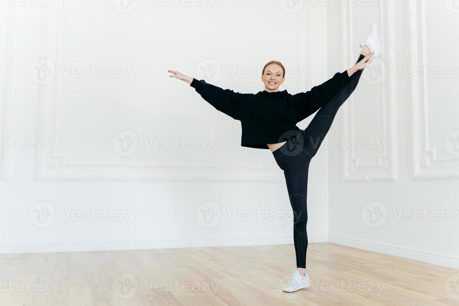 une femme sportive souriante se tient sur une jambe dans un centre de remise en forme, fait preuve d'une belle résistance physique, a une silhouette mince, vêtue d'un sweat à capuche et de leggings noirs, porte des baskets blanches. les gens et l'équilibre photo