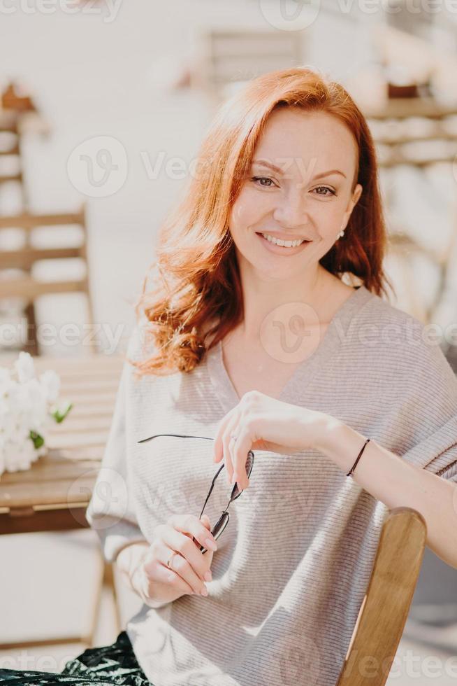 photo d'une belle femme au gingembre avec une expression satisfaite, tient des lunettes de soleil, a du temps libre, s'assoit dans un café extérieur, a une conversation agréable avec un interlocuteur, profite d'une bonne journée ensoleillée. concept de personnes et de repos