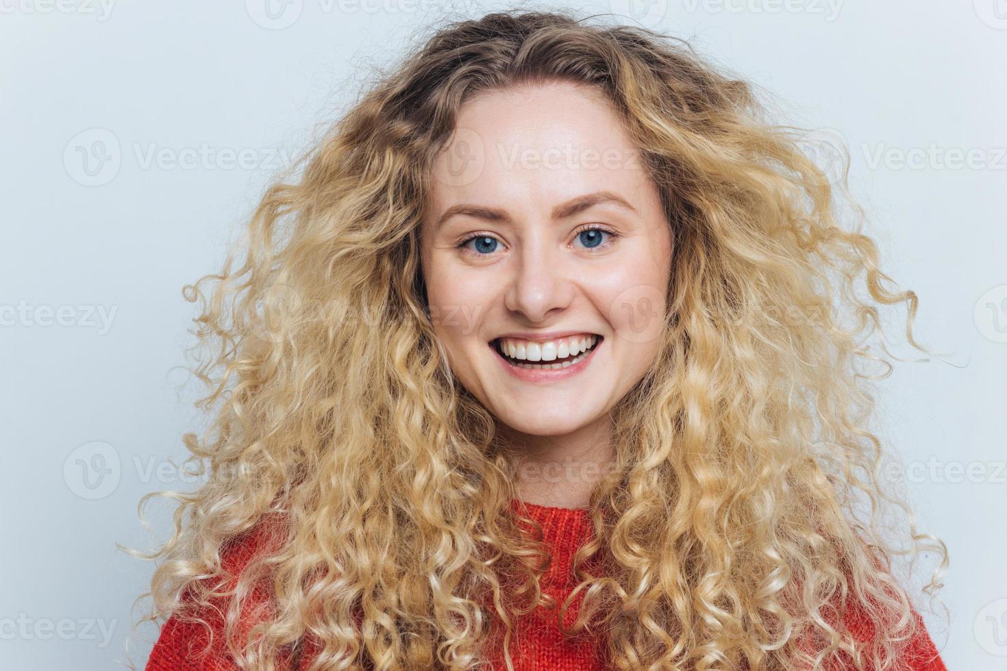 gros plan d'une adorable femme heureuse avec des cheveux bouclés, une peau saine, un large sourire et des dents blanches parfaites, heureuse de passer du temps libre avec les meilleurs amis en drôle de compagnie, pose contre le mur blanc du studio photo