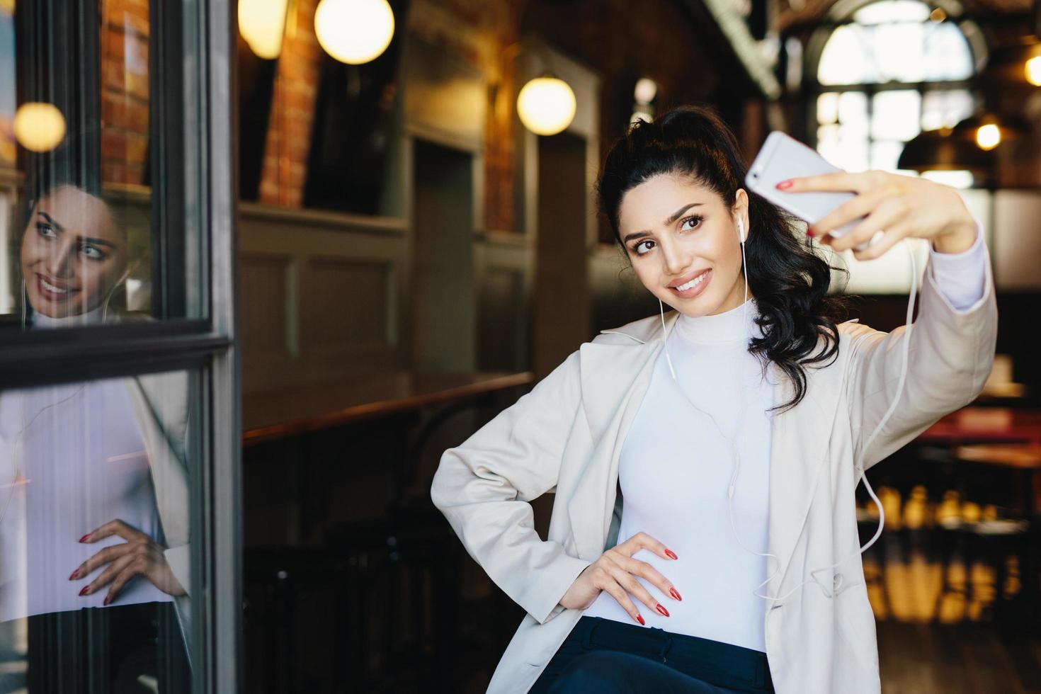 joyeuse femme brune en vêtements blancs élégants tenant un téléphone portable générique faisant sefie sur fond de café confortable. jeune femme à la mode faisant une photo d'elle-même en s'amusant. technologie moderne