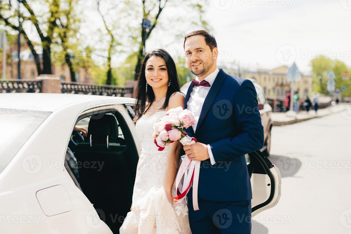 un joli couple marié se tient l'un à côté de l'autre près de la voiture, va au restaurant pour célébrer son mariage, faire la fête, inviter des amis et des parents. les jeunes mariés posent en plein air après l'enregistrement du mariage photo