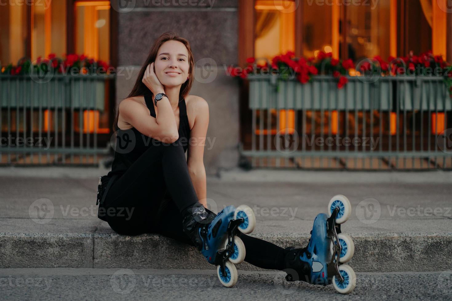 photo horizontale d'une jeune femme active prend une pause tout en faisant du roller dans la rue vêtue de vêtements de sport noirs se sent satisfaite des sourires pose agréablement en plein air. mode de vie d'été et concept de repos