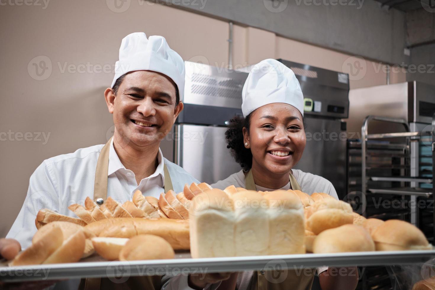 portrait de chefs professionnels en uniforme blanc regardant la caméra avec un sourire joyeux et fier avec un plateau de pain dans la cuisine. un ami et partenaire des aliments de boulangerie et de l'occupation quotidienne de la boulangerie fraîche. photo