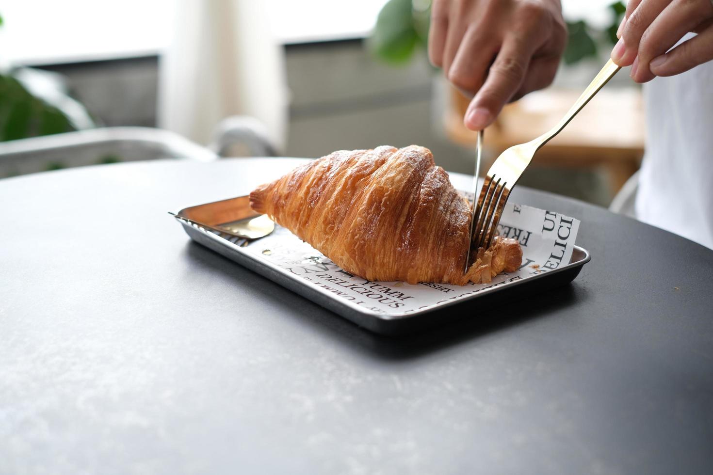 gros plan de la main utilise une fourchette et un couteau pour couper un morceau de croissant à manger pour le petit-déjeuner sur une table en bois au café. photo