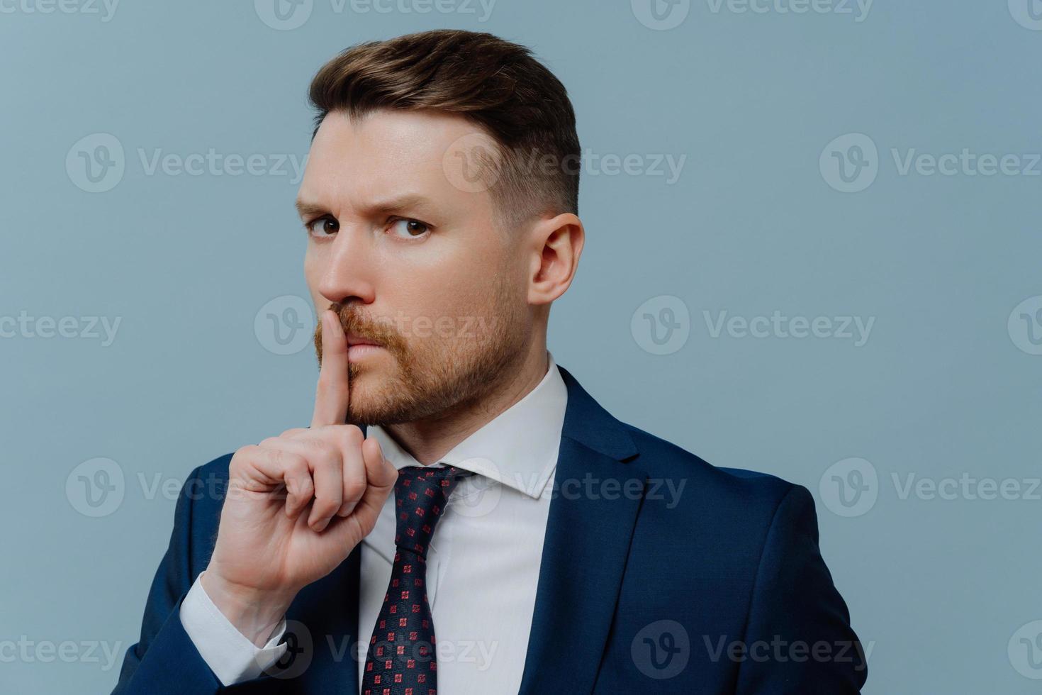 un employé de bureau masculin prospère barbu sérieux vêtu de vêtements formels fait un geste silencieux demande de garder le silence exige le silence avec une expression stricte isolée sur fond bleu. tabou chut chut photo