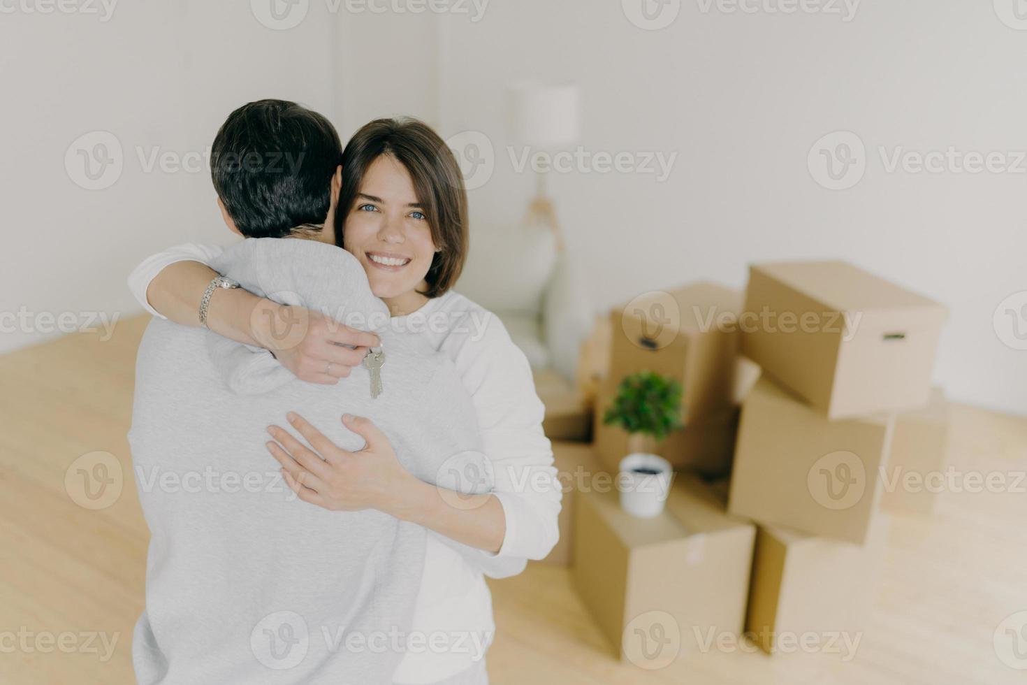 photo intérieure d'un charmant couple familial s'embrasse avec amour, tient les clés du nouvel appartement, emménage dans son propre appartement, pose dans un salon lumineux vide avec lampadaire et tas de nombreux conteneurs en carton