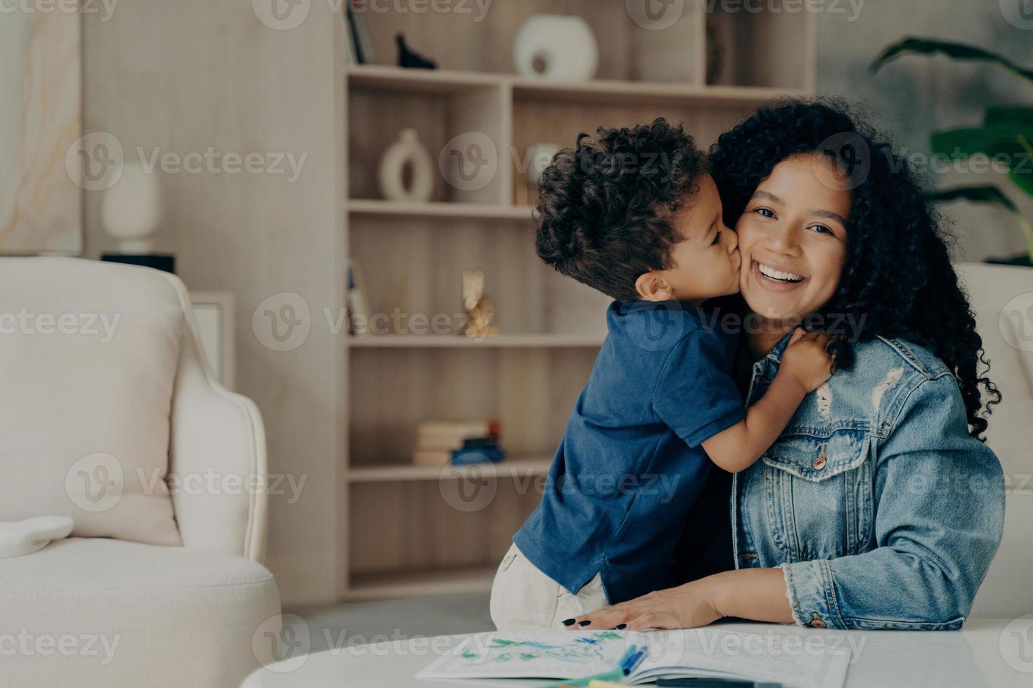 portrait d'un garçon afro-américain mignon en t-shirt polo bleu étreignant sa mère et l'embrassant sur la joue photo