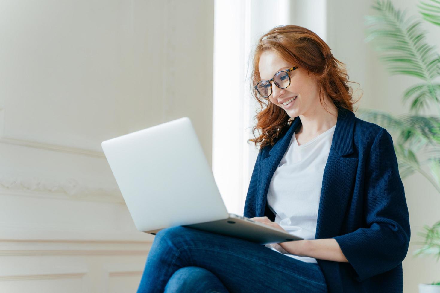 une employeur professionnelle aime le processus de travail, s'assoit les jambes croisées avec un ordinateur portable, discute en ligne, a un regard positif, a les cheveux bouclés rouges, porte des vêtements élégants et élégants. les gens et la technologie photo