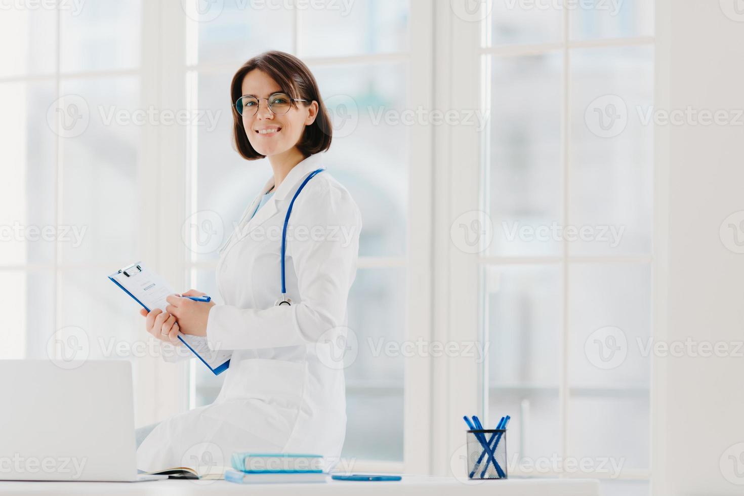 une femme médecin qualifiée écrit dans le presse-papiers, pose sur le lieu de travail avec un ordinateur portable. médecin de famille professionnel prêt à vous donner des conseils pour prendre soin de votre santé, porte un uniforme médical. photo