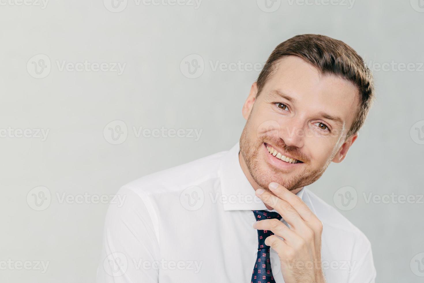 photo horizontale d'un homme gai avec chaume, tient le menton, regarde positivement la caméra, vêtu d'une chemise formelle avec cravate, écoute quelque chose d'agréable, isolé sur fond blanc. homme employé