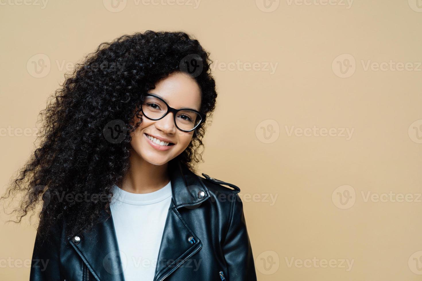 photo horizontale d'une femme heureuse en bonne santé avec des cheveux bouclés naturels, un sourire agréable, aime le temps libre et faire du shopping le week-end, porte des lunettes transparentes, une veste en cuir, isolée sur fond beige