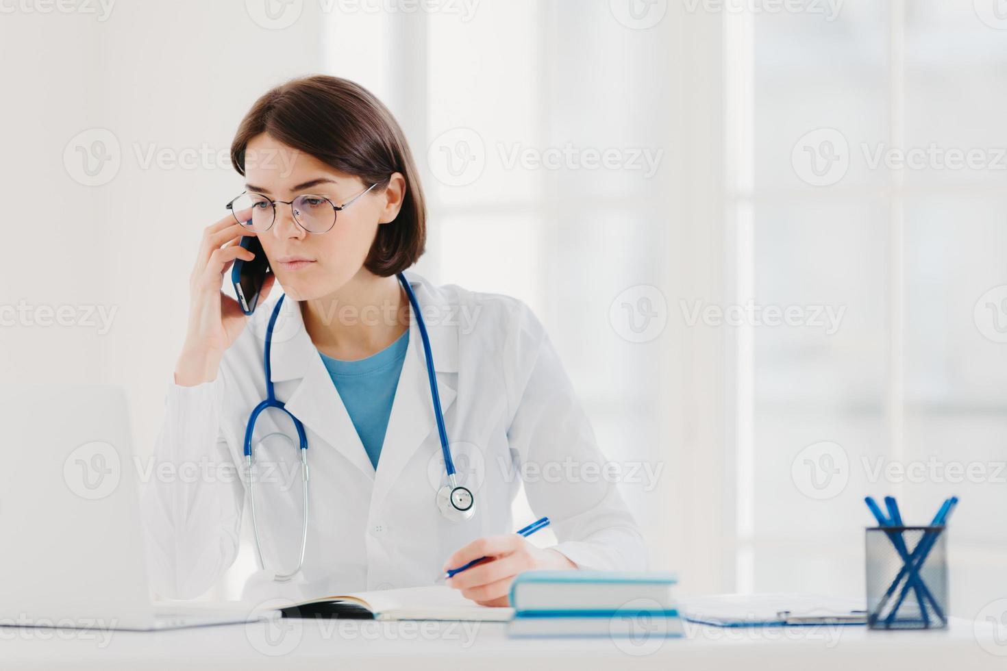 personnel de soins de santé, concept de médecine. femme médecin brune sérieuse concentrée sur un ordinateur portable moderne, réécrit les informations nécessaires, parle sur un téléphone portable, appelle quelqu'un, a un regard sérieux photo