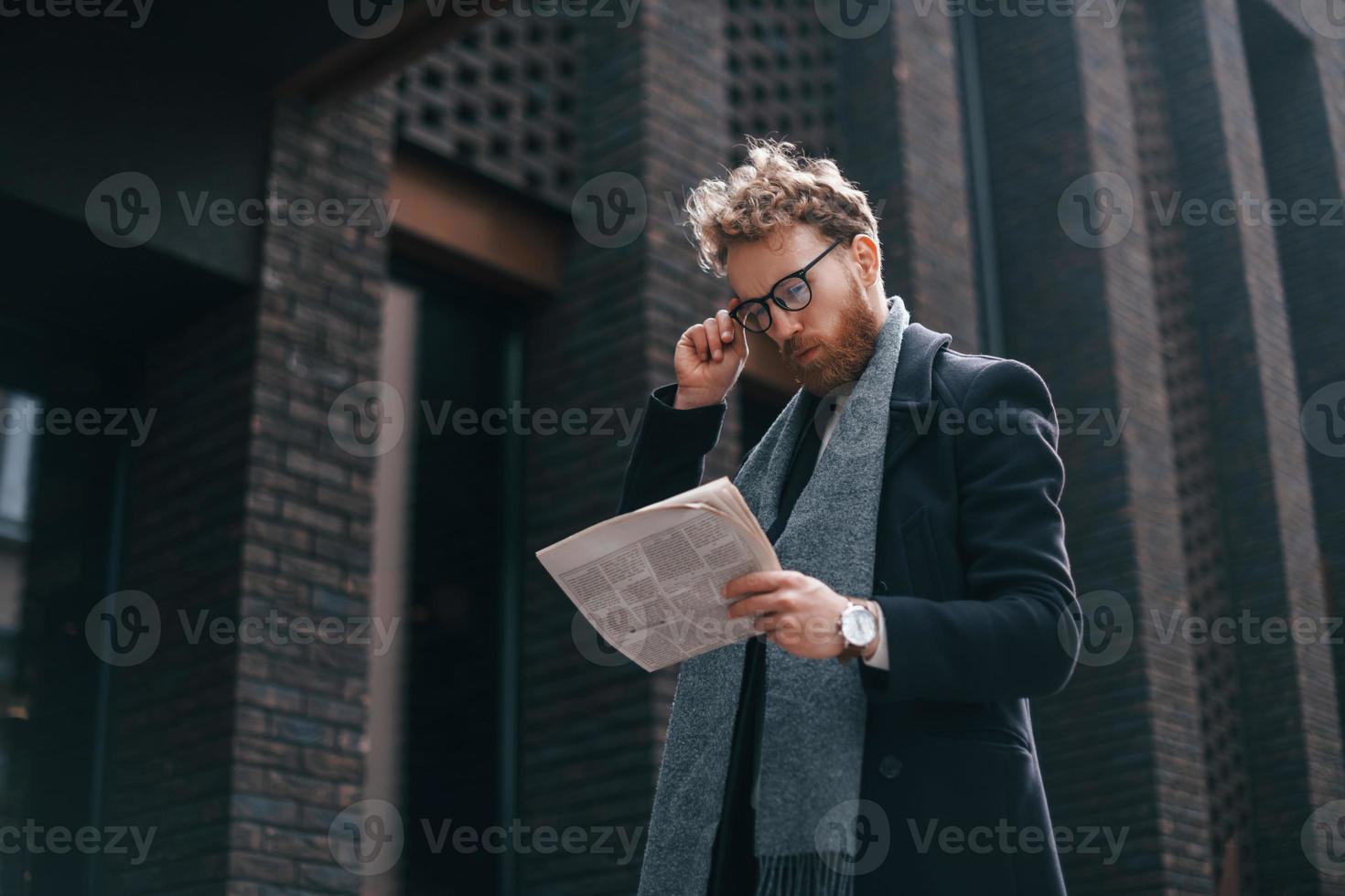 avec un journal dans les mains. homme élégant avec barbe et lunettes est à l'extérieur près du bâtiment photo