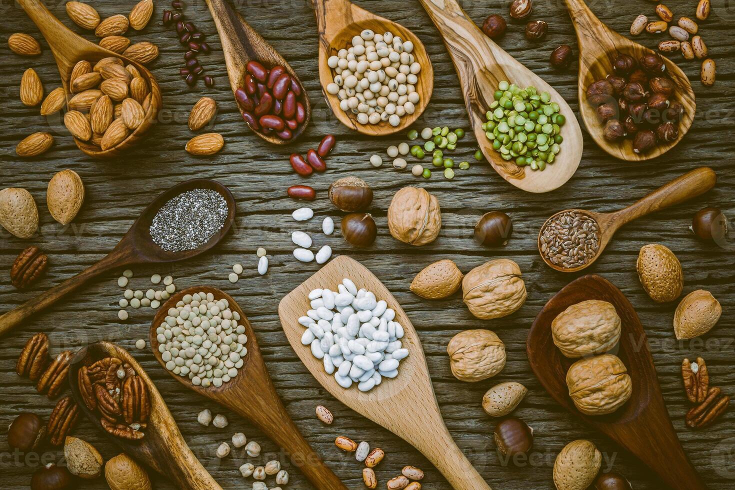 diverses légumineuses et différents types de coquilles de noix dans des cuillères. cerneaux de noix, noisettes, amandes, pinto brun, graines de soja, graines de lin, chia, pois chiches, haricots rouges et noix de pécan sur une table en bois minable. photo