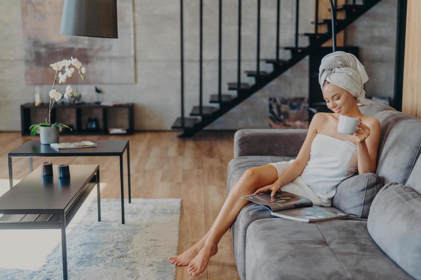 femme détendue avec des jambes fines, une peau saine, aime boire du thé, s'assoit sur un canapé confortable, enveloppée dans une serviette de bain, pose contre un intérieur confortable. personnes, beauté, détente, concept de style de vie photo