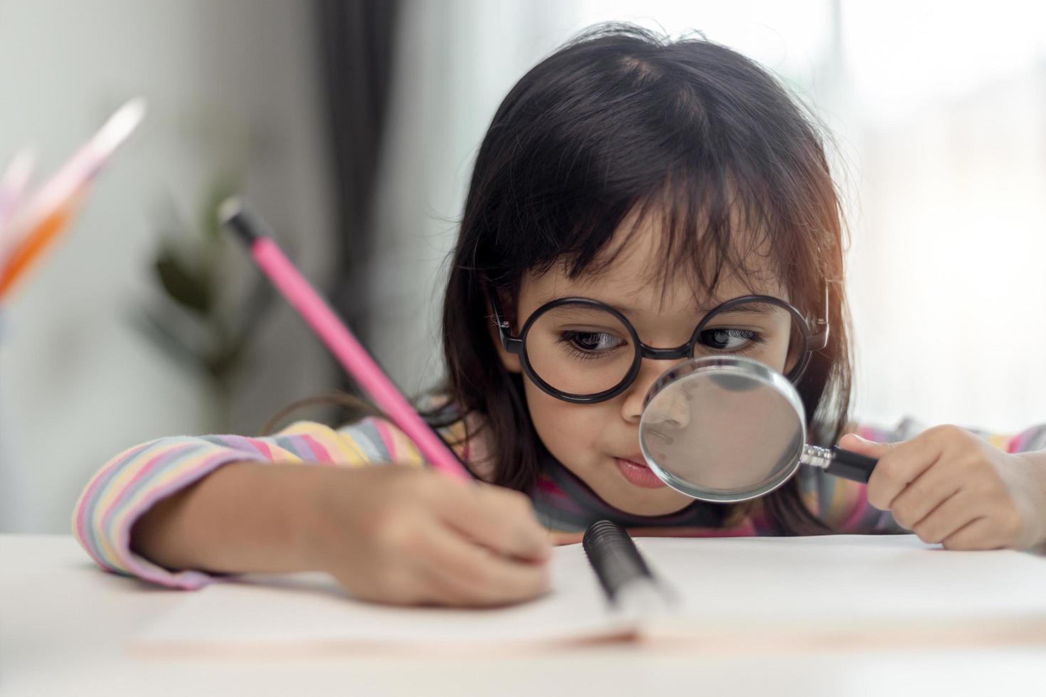 étudiante petite fille asiatique écrivant avec une loupe en regardant la caméra. adorables enfants assis dans le salon à la maison. concept d'activité familiale. photo