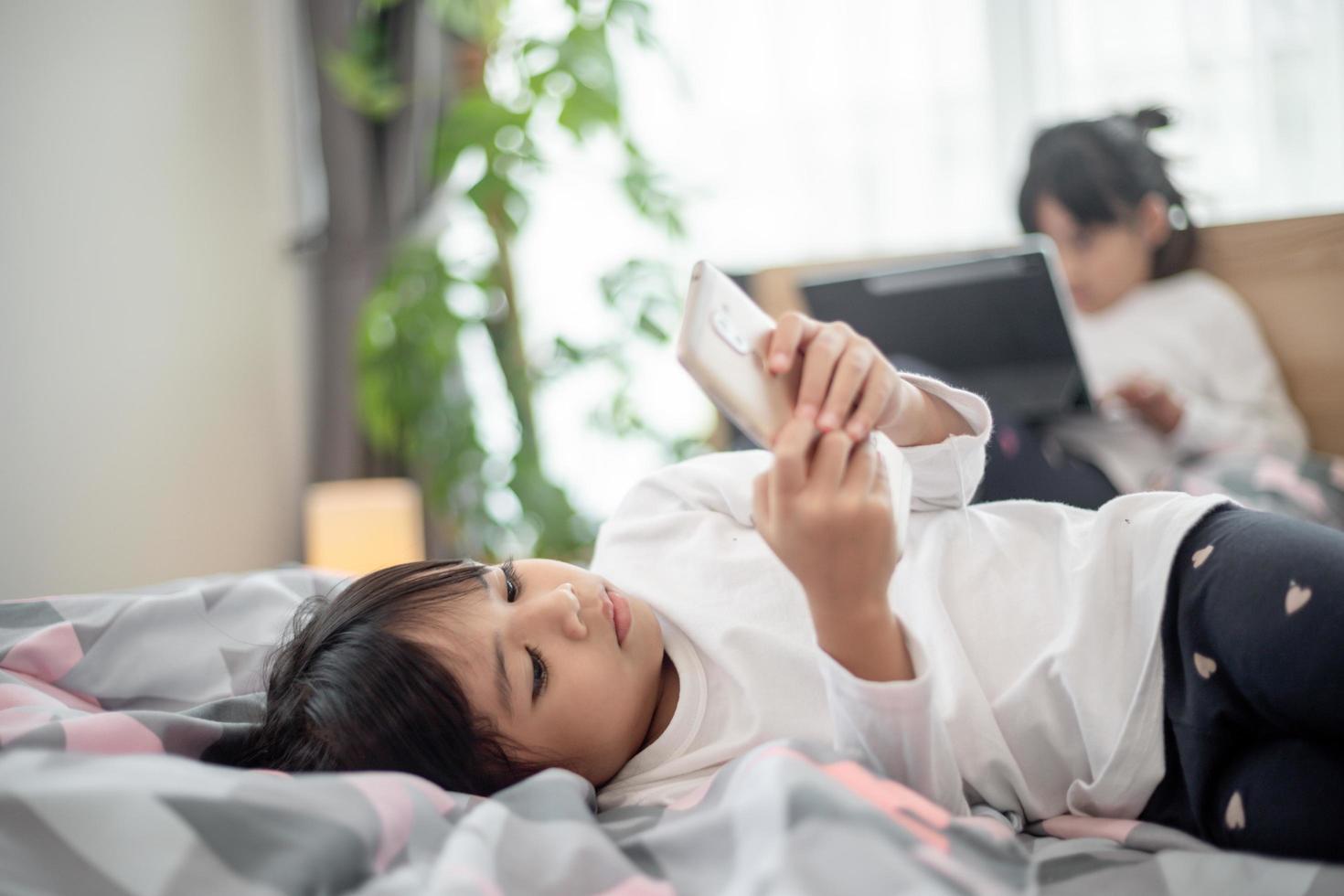 jolie petite fille regardant une vidéo sur un smartphone avec un visage souriant seul sur le lit, enfant utilisant un téléphone portable avec un visage heureux à la maison. rester à la maison quarantaine coronavirus covid-19 prévention de la pandémie. photo