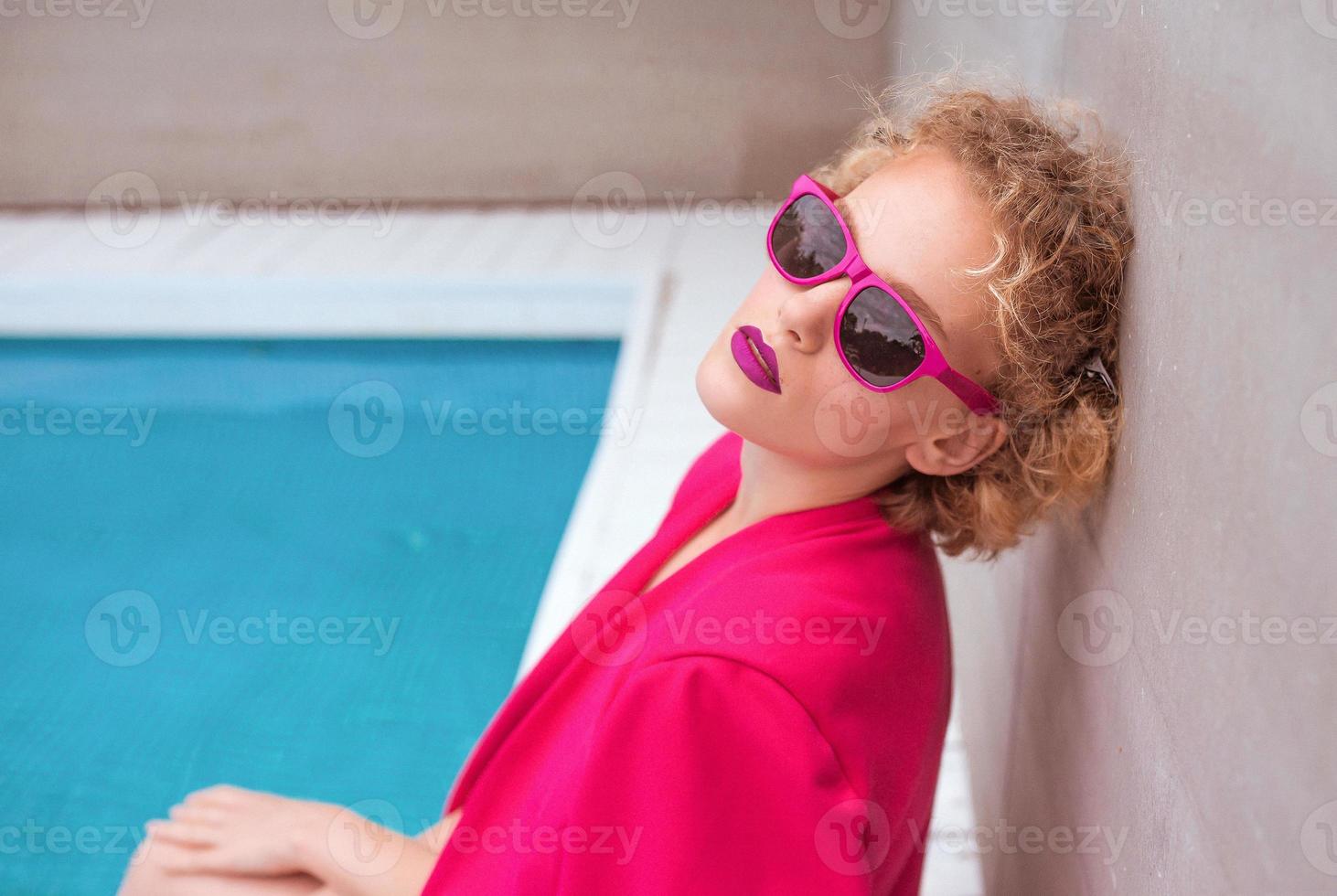 portrait d'une merveilleuse jeune femme bouclée rousse élégante assise en veste fuchsia, lunettes de soleil élégantes et sous-vêtements noirs au bord de la piscine. photo