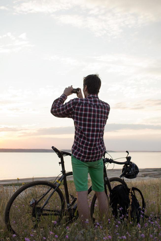 silhouette d'un homme avec un vélo de route de randonnée regardant et faisant une photo du coucher du soleil dans le lac sur un téléphone portable