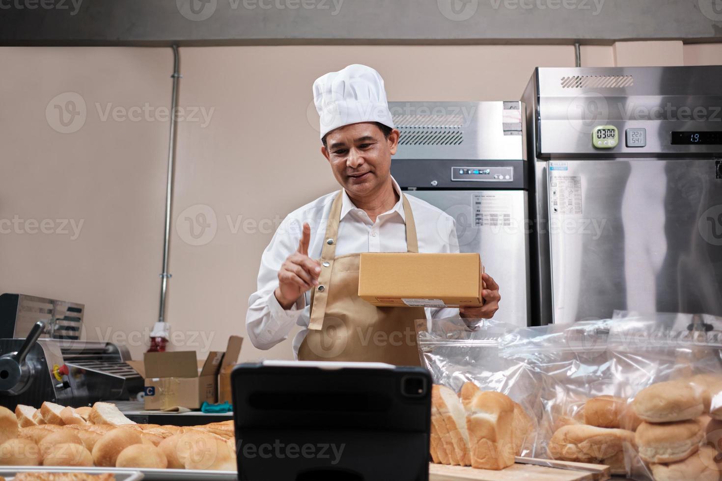chef masculin asiatique senior en tablier, blogueur de petite entreprise en direct via la technologie d'application mobile, promotion en ligne des ventes de boulangerie, pain frais, pâtisserie et cuisine commerciale dans la cuisine. photo