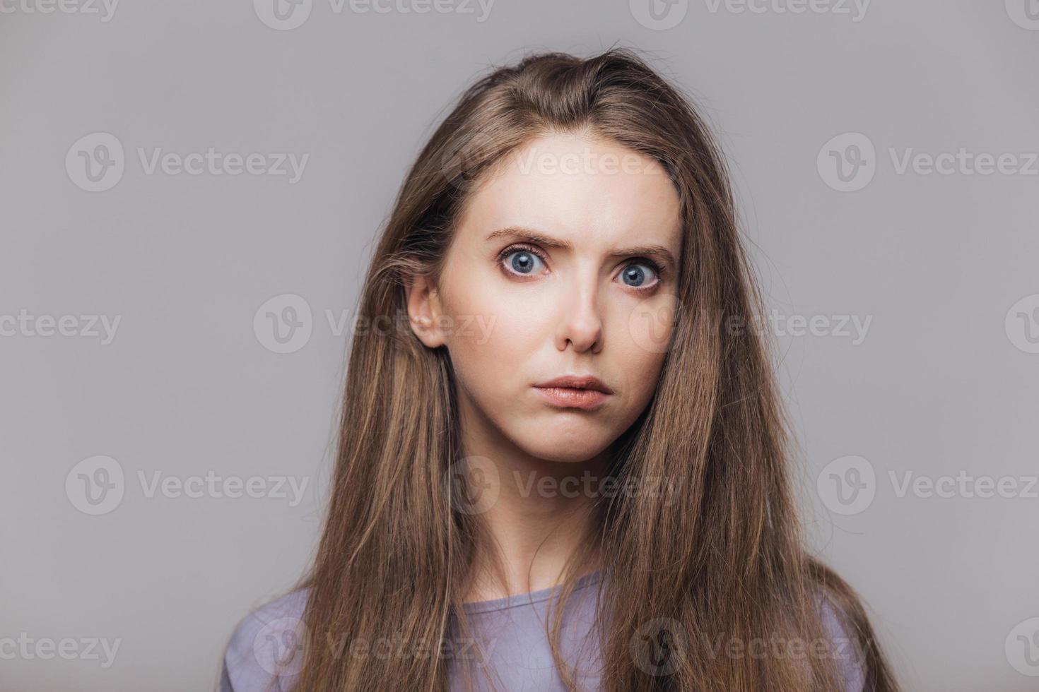 photo de tête d'une belle femme aux yeux bleus et à la peau pure, regarde sérieusement et avec colère la caméra, a les cheveux raides foncés, isolée sur fond gris. photo horizontale d'une belle jeune femme à l'intérieur