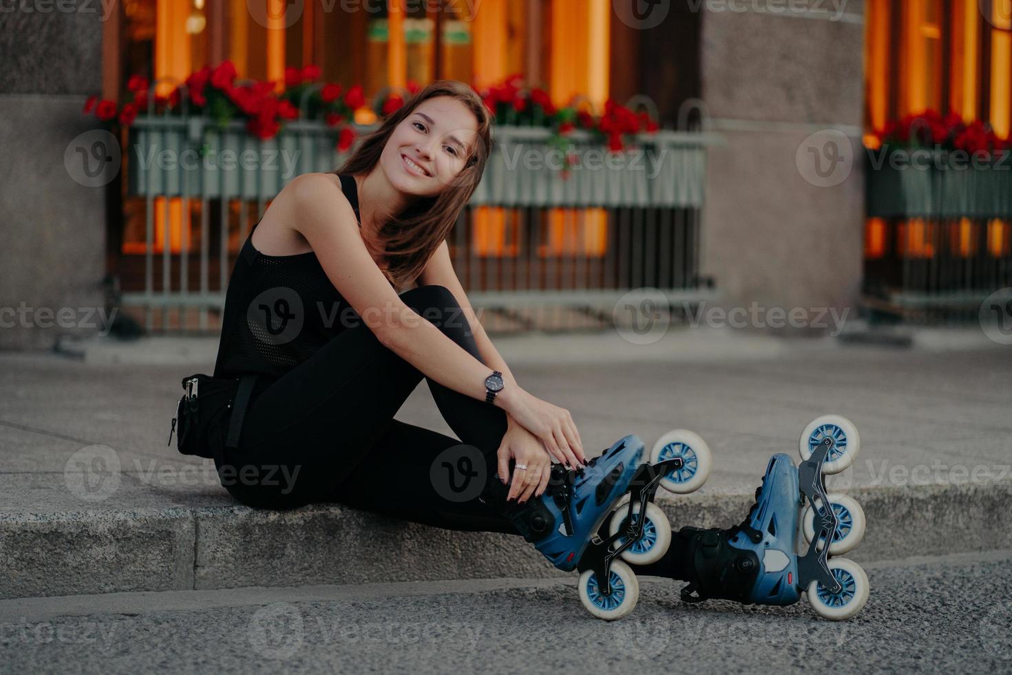 une jeune femme européenne à l'allure agréable porte des patins à roulettes prend une pause après avoir roulé pose en plein air vêtue de vêtements de sport noirs a un sourire heureux sur le visage. loisirs loisirs et concept de style de vie photo