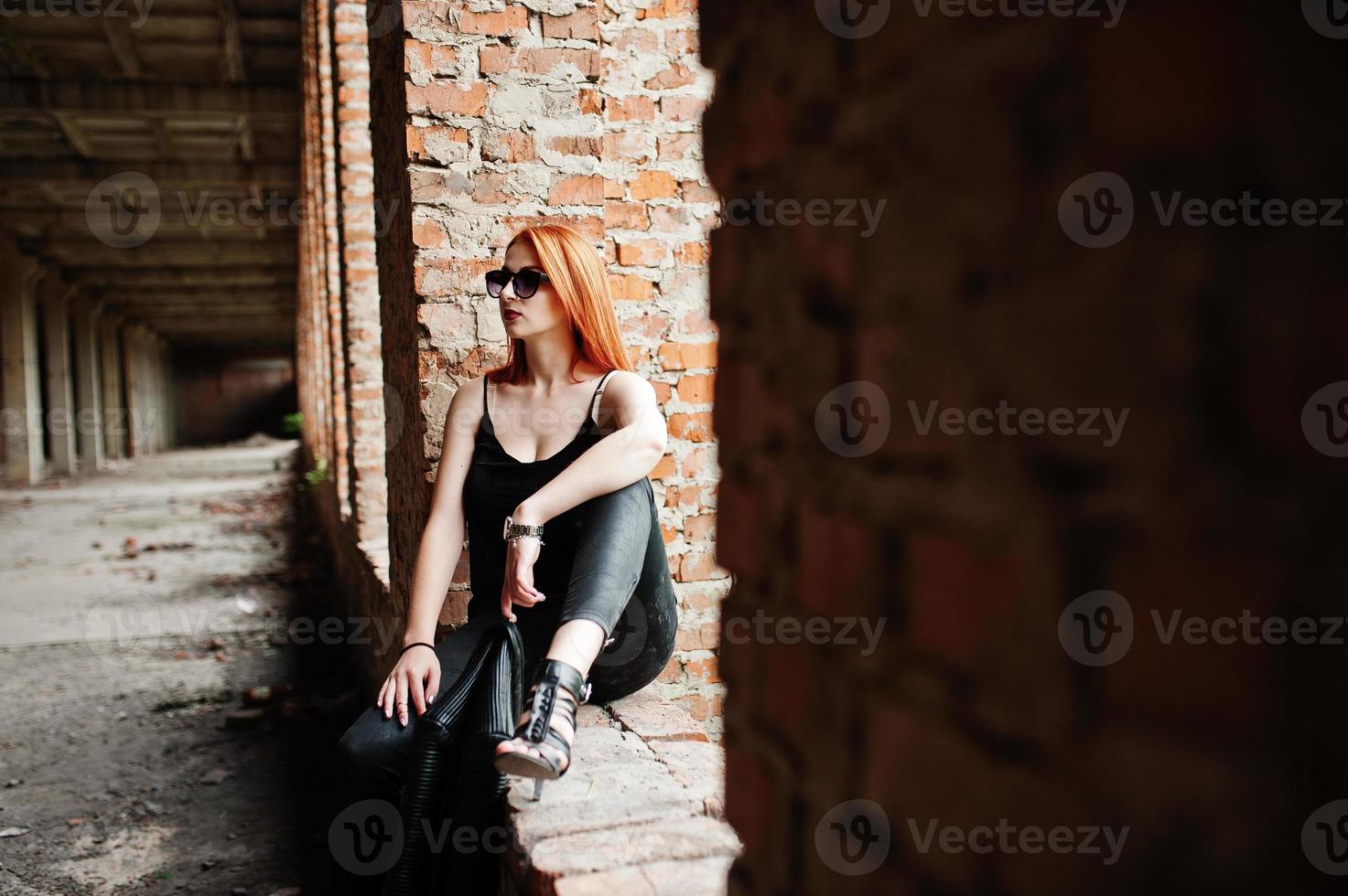 fille élégante aux cheveux rouges portant des lunettes de soleil en noir, contre un endroit abandonné avec des murs de briques. photo