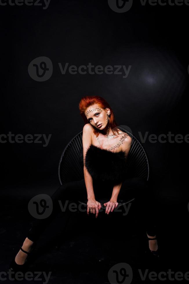 modèle de mode fille aux cheveux rouges avec à l'origine maquillage comme prédateur de léopard isolé sur noir. portrait en studio sur chaise. photo