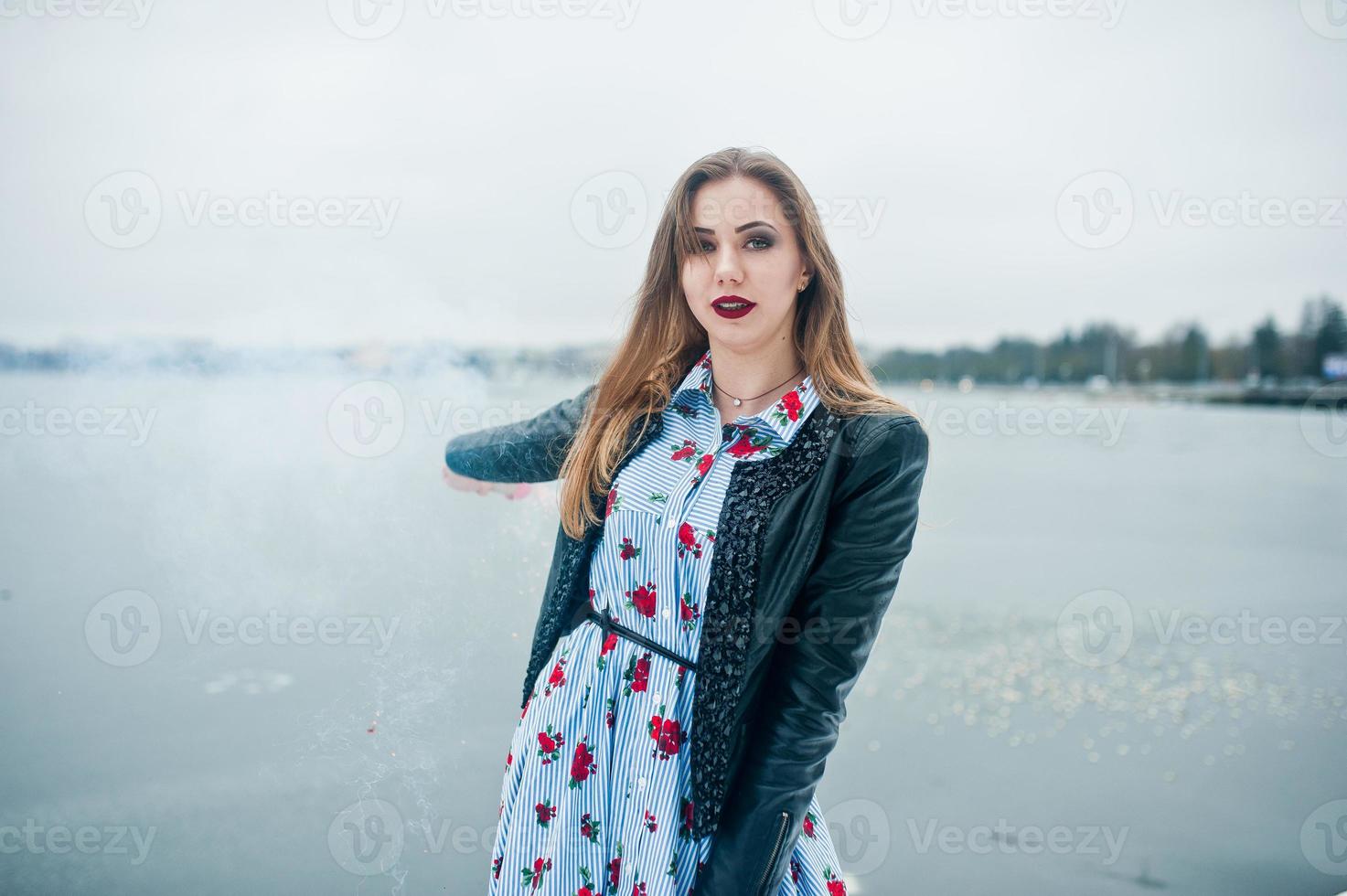 une fille élégante en veste de cuir tient une fusée de fumée rose le jour de l'hiver contre un lac gelé. photo