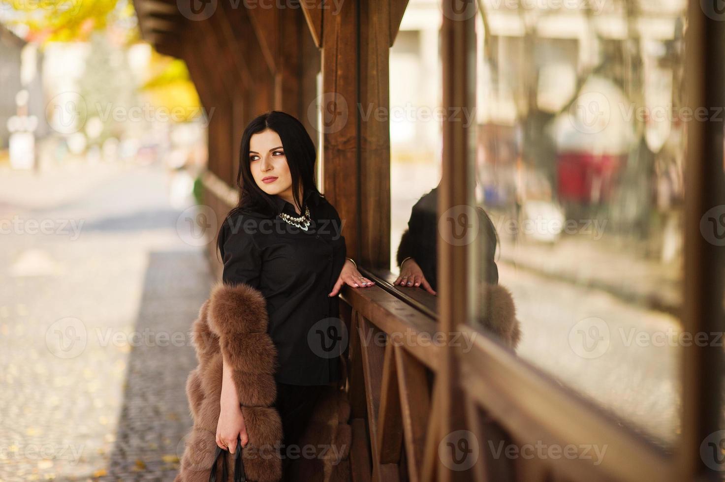 photo de mode en plein air d'une magnifique femme sensuelle aux cheveux noirs dans des vêtements élégants et un manteau de fourrure luxueux contre une terrasse en bois à la ville d'automne.