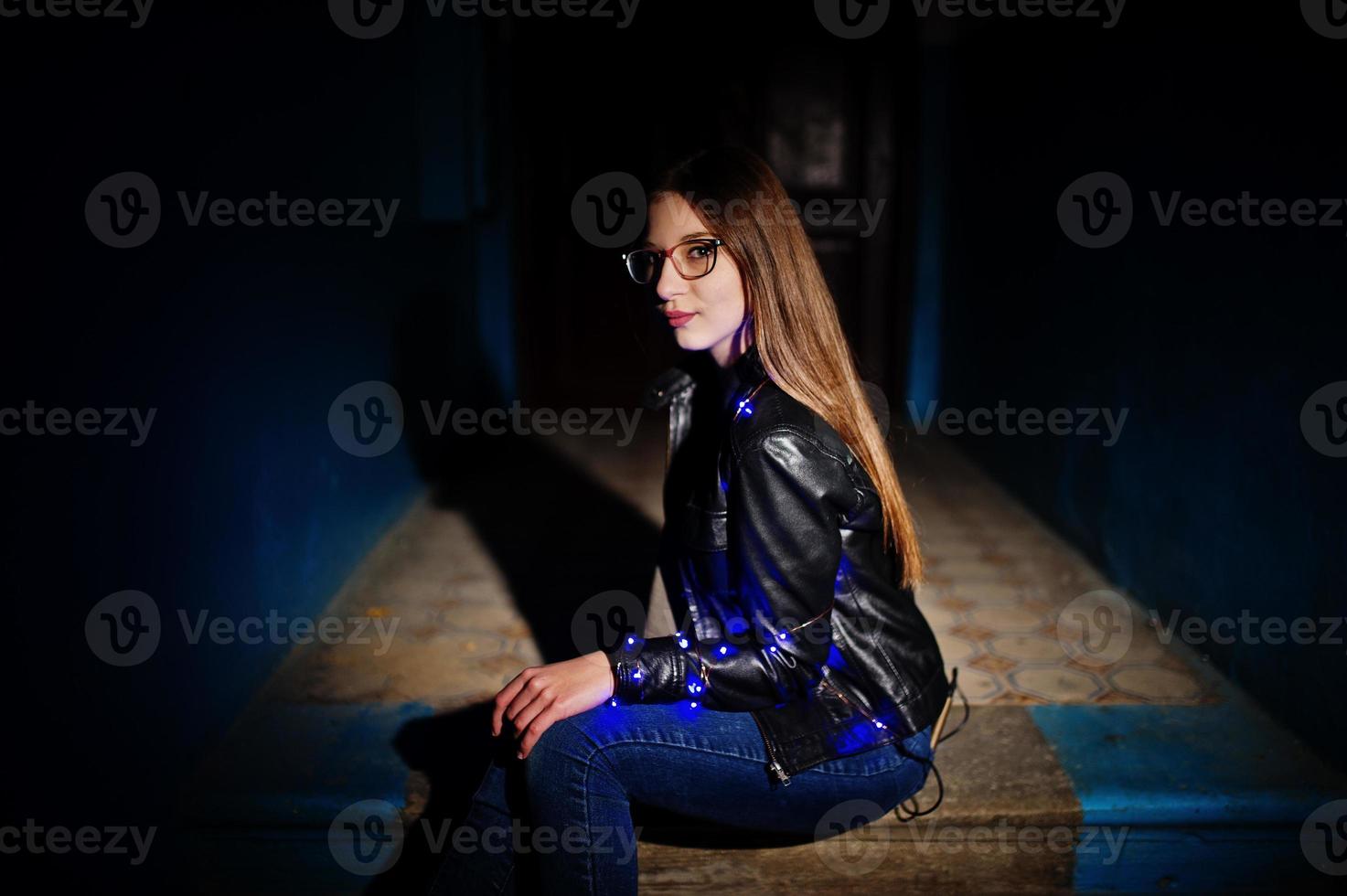 portrait de nuit d'une fille modèle portant des lunettes, un jean et une veste en cuir, avec une guirlande bleue sur elle. photo