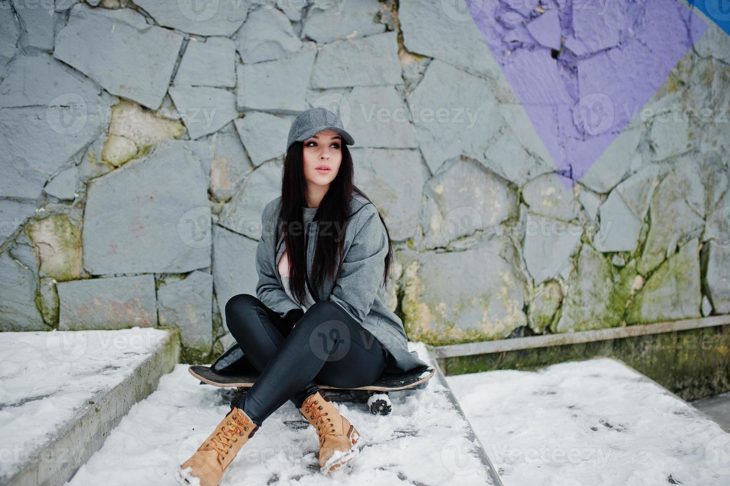 fille brune élégante en casquette grise, style de rue décontracté avec planche à roulettes le jour de l'hiver contre un mur coloré. photo