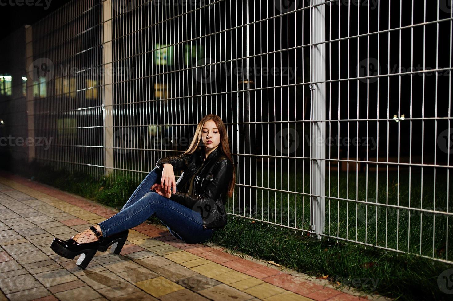 portrait de nuit d'une fille modèle portant un jean et une veste en cuir contre une clôture en fer. photo