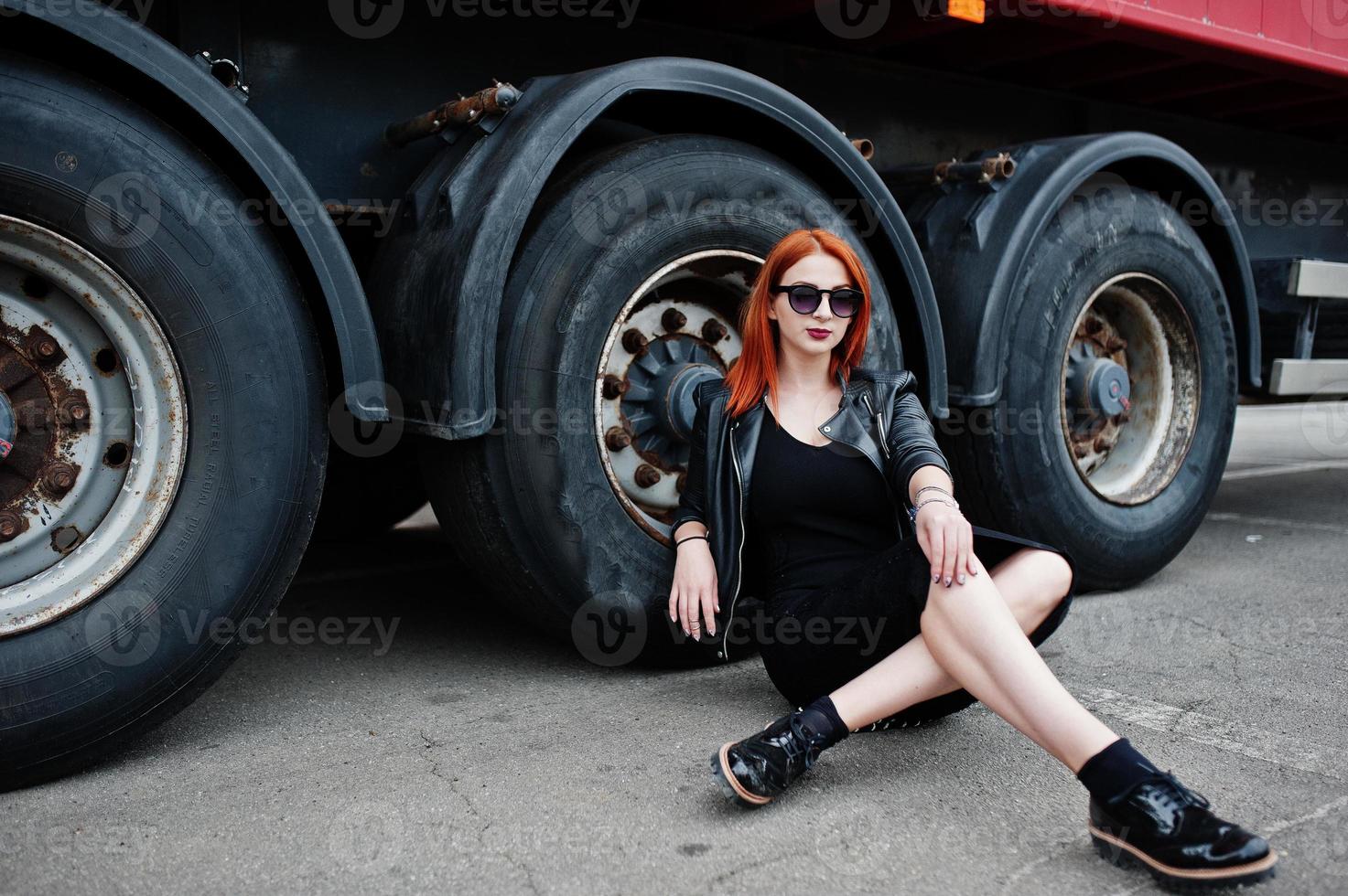 fille élégante aux cheveux rouges en noir, assise contre de grandes roues de camion. photo