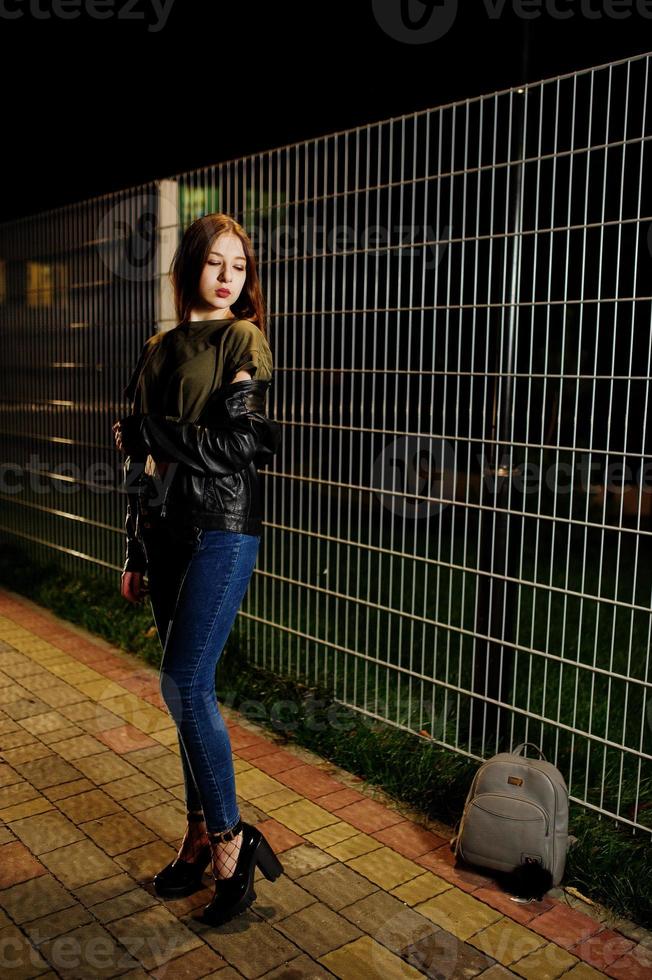 portrait de nuit d'une fille modèle portant un jean et une veste en cuir contre une clôture en fer. photo