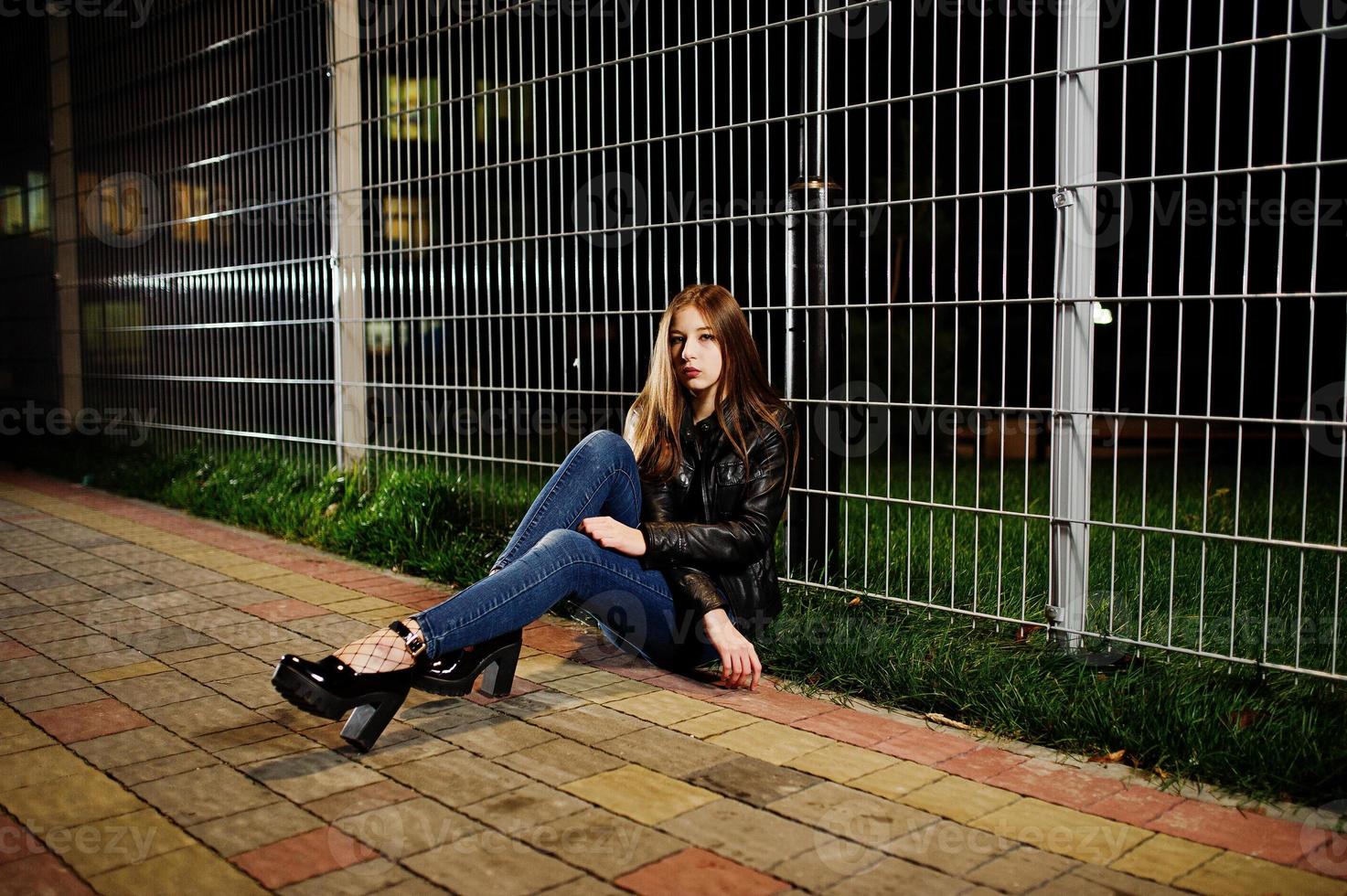 portrait de nuit d'une fille modèle portant un jean et une veste en cuir contre une clôture en fer. photo