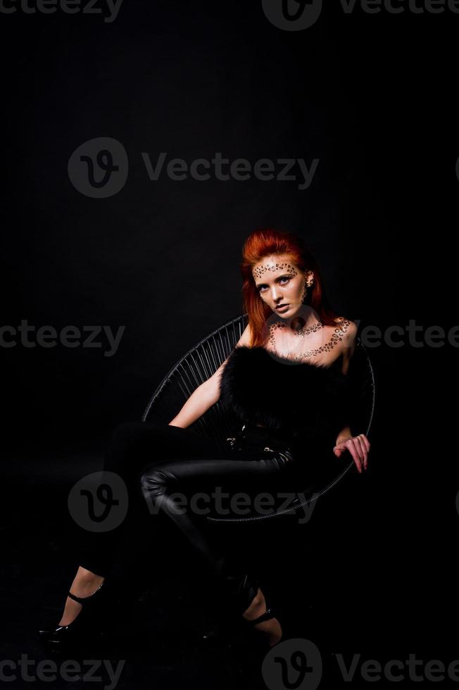 modèle de mode fille aux cheveux rouges avec à l'origine maquillage comme prédateur de léopard isolé sur noir. portrait en studio sur chaise. photo