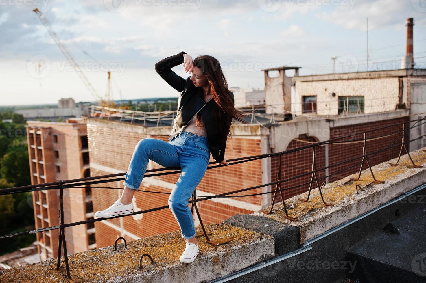 portrait d'une magnifique jeune femme en veste de cuir noir, jeans et baskets assis sur des mains courantes sur le toit avec vue pittoresque sur un parc. photo