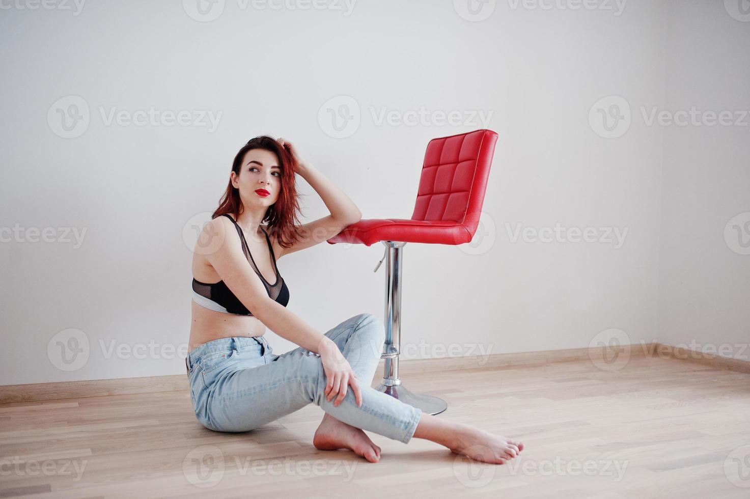 fille aux cheveux rouges avec un gros buste sur un soutien-gorge noir et un jean assis près d'une chaise rouge contre un mur blanc dans une pièce vide. photo