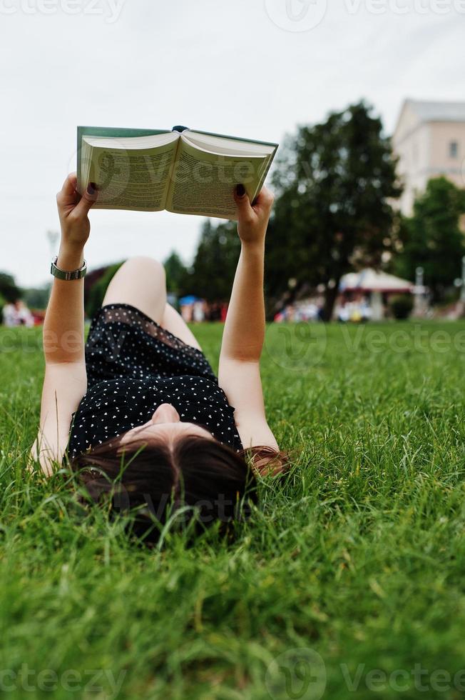 portrait d'une belle femme en robe noire à pois allongée sur l'herbe et lisant un livre dans le parc. photo