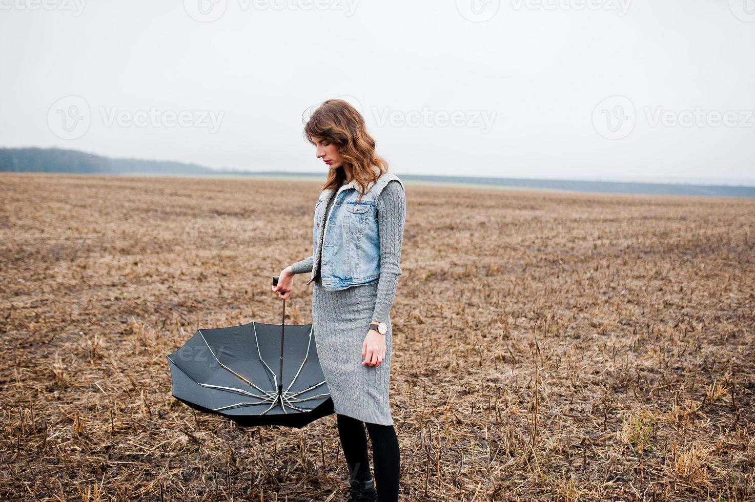 portrait de jeune fille bouclée brune en veste jeans avec parapluie noir sur le terrain. photo