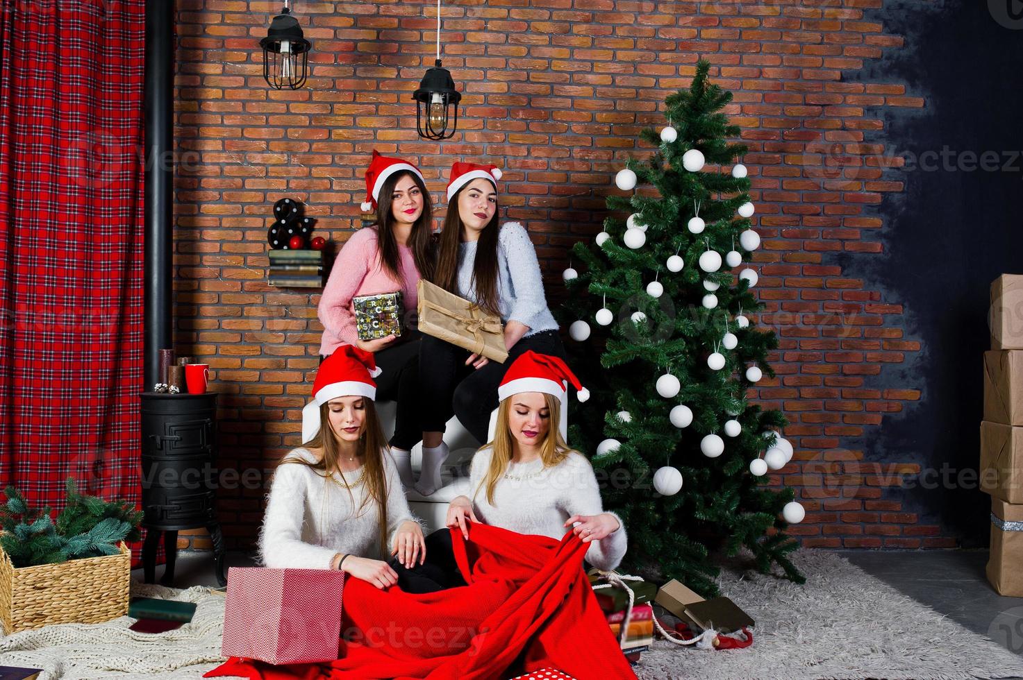 quatre amies mignonnes que les filles portent sur des chandails chauds, des pantalons noirs et des chapeaux de père noël contre l'arbre du nouvel an avec une décoration de noël au studio. photo