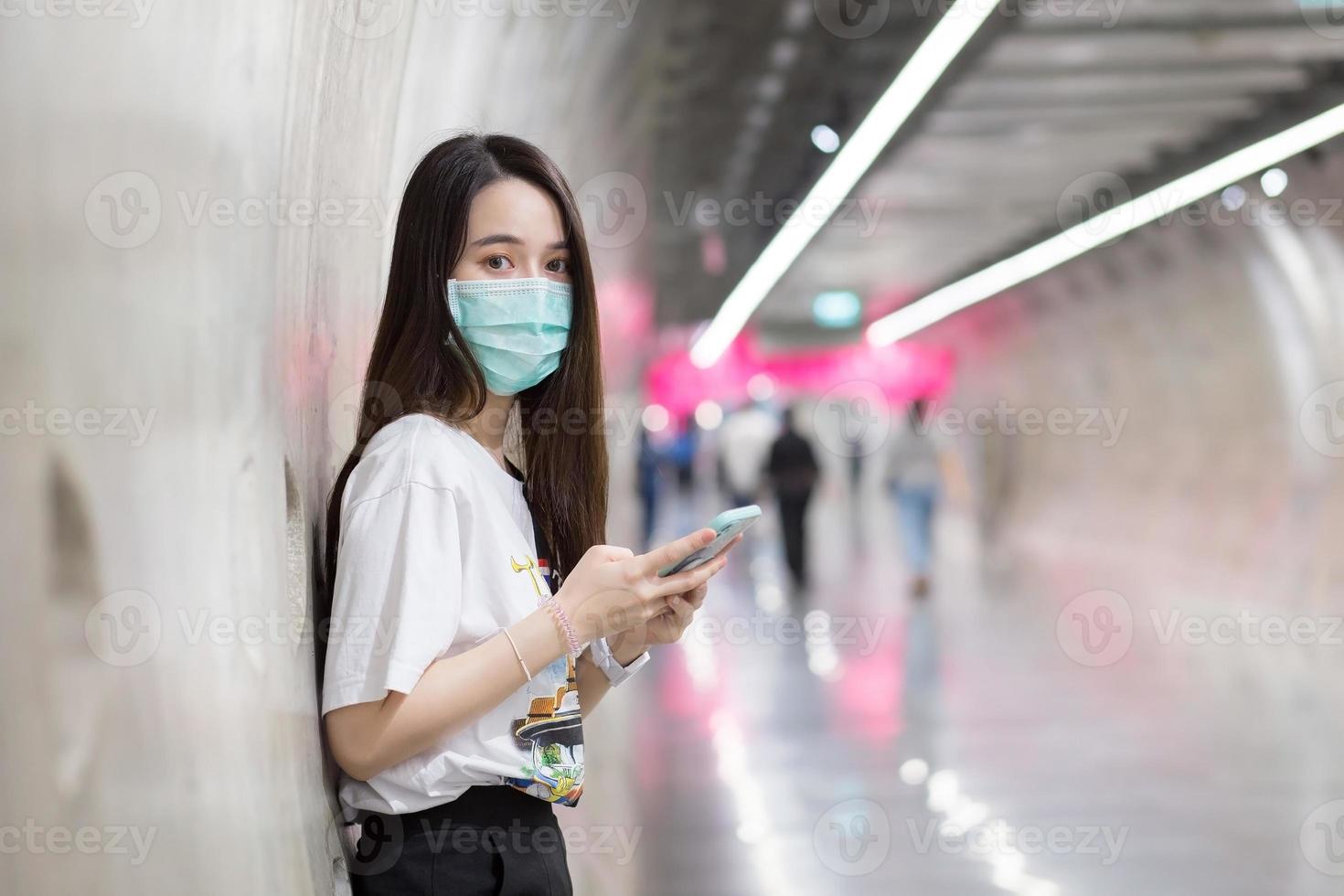 une jeune femme asiatique se tient dans le tunnel du métro et porte un masque facial comme nouveau mode de vie normal et de soins de santé tout en tenant un smartphone dans sa main. photo