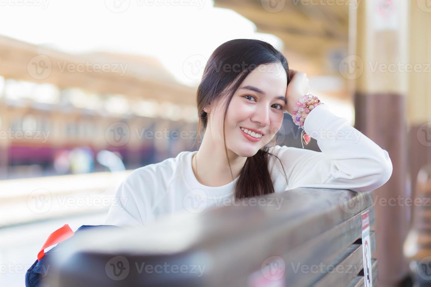 une belle femme asiatique dans une chemise blanche à manches longues et un chapeau est assise dans la gare en attendant l'arrivée du train. photo