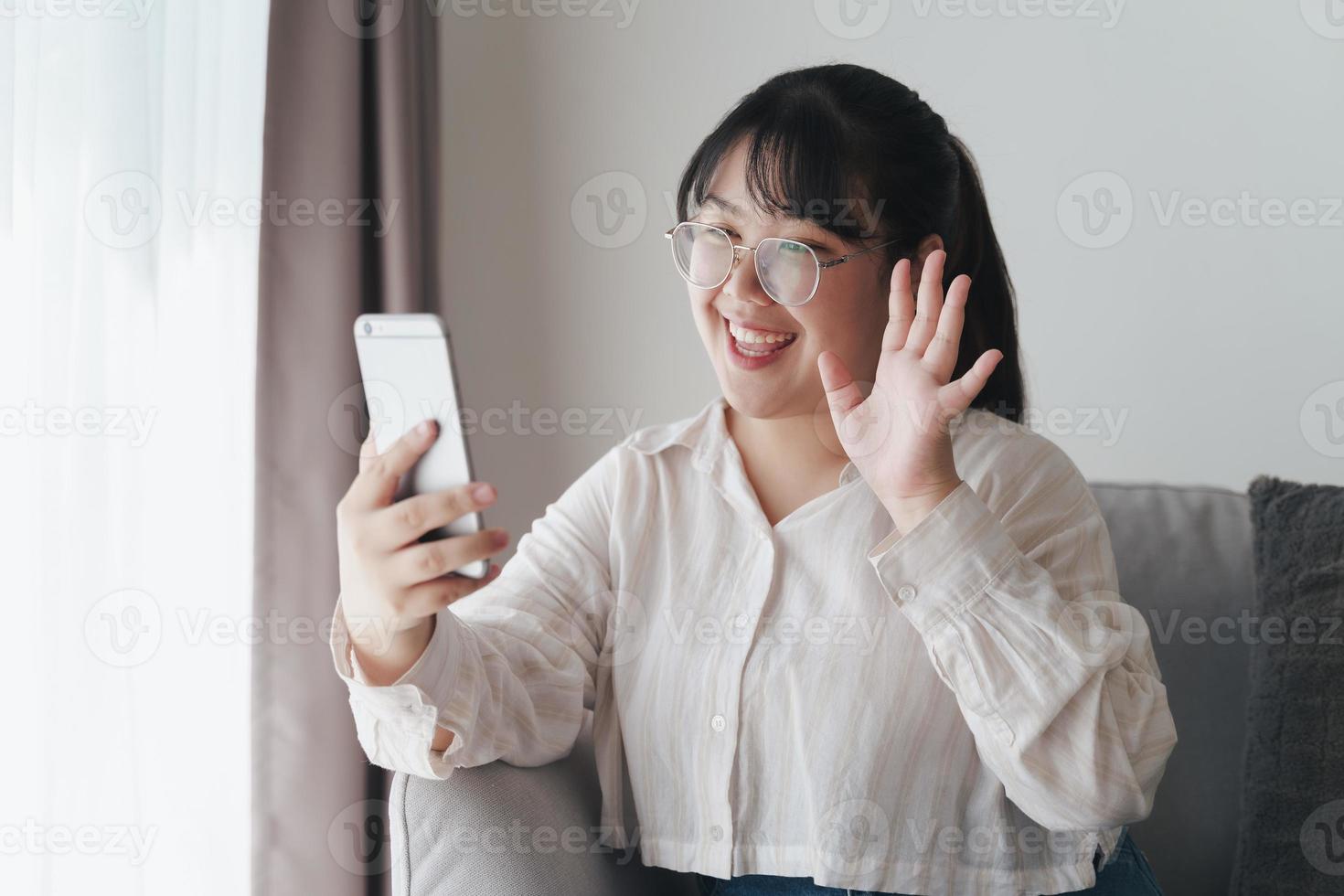 jeune femme asiatique utilisant un smartphone pour une visioconférence en ligne en agitant la main faisant un geste de bonjour sur le canapé dans le salon. photo