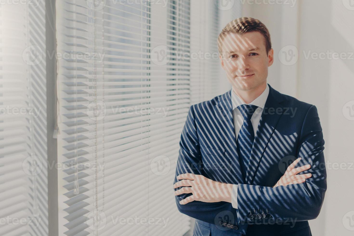 un homme d'affaires attrayant se tient les bras croisés près de la fenêtre du bureau avec jalousie, vêtu de vêtements élégants de luxe, se repose après avoir rencontré son partenaire, a une expression satisfaite. style de vie d'affaires photo