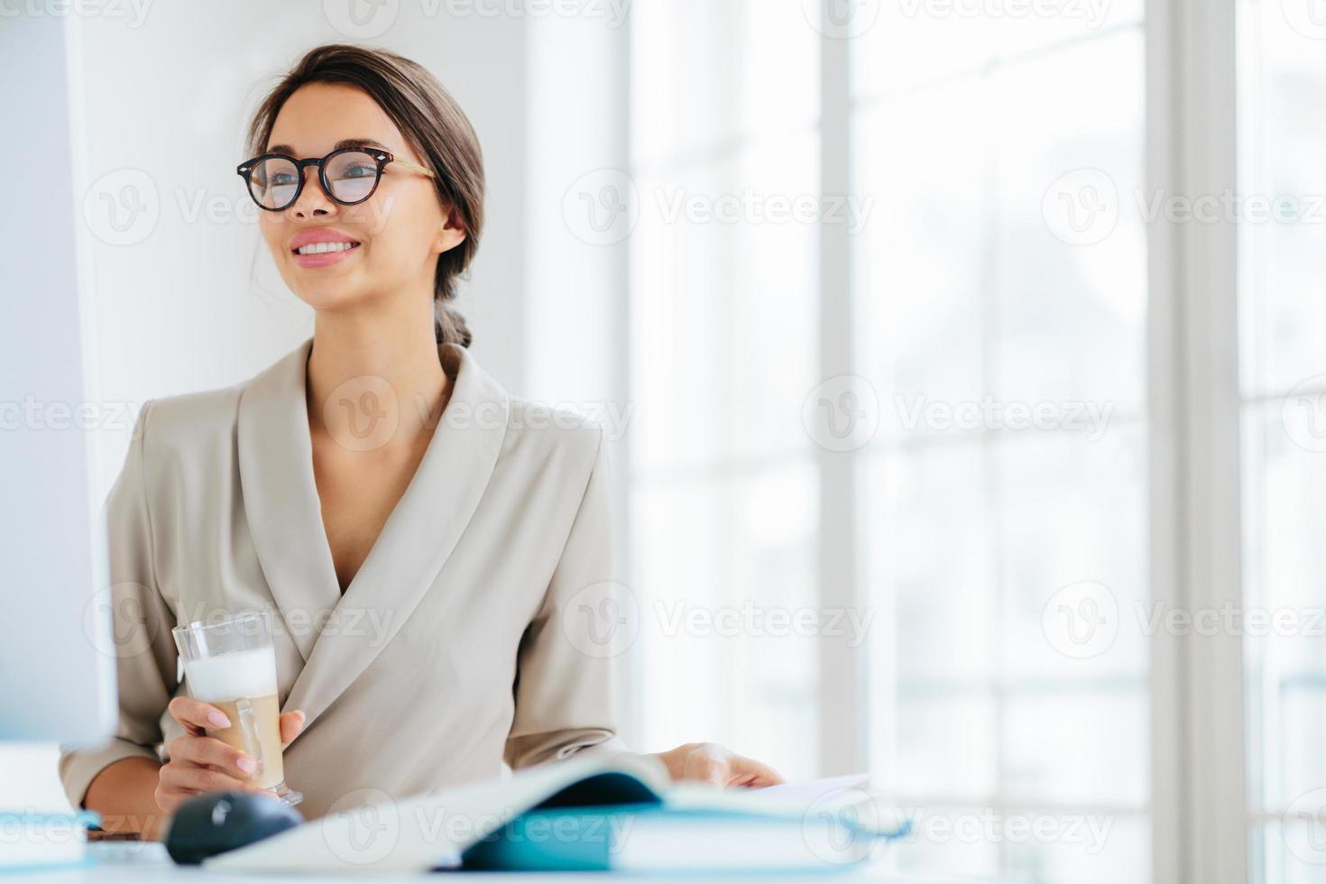 une femme positive travaille au bureau, s'assoit au bureau, se concentre sur le moniteur, boit du milkshake frais, sourit et aime travailler, pose contre l'intérieur du bureau pense à l'organisation d'un événement professionnel photo