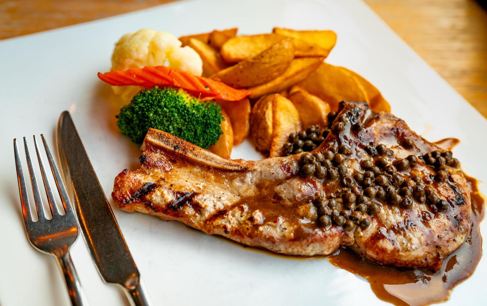 steak nappé de sauce et de poivre servi avec pommes de terre frites et carottes, chou-fleur et brocoli cuits à la vapeur. steak sur plaque blanche avec fourchette et couteau en acier inoxydable posés sur une table en bois. bifteck de tomahawk. photo