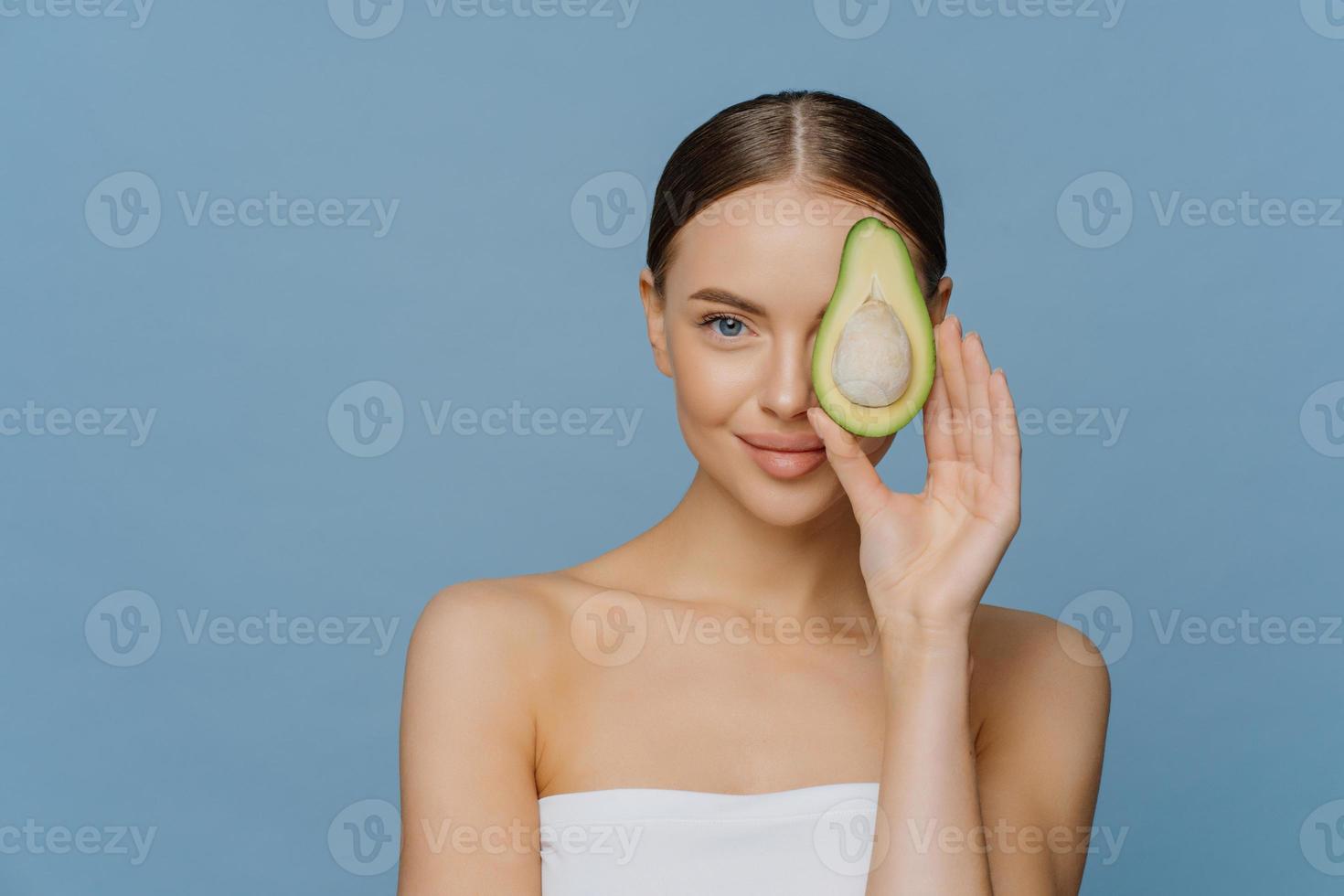 concept de soins du visage. charmante femme tendre aux cheveux noirs peignés a une peau brillante couvre les yeux avec la moitié de l'avocat obtient des huiles essentielles pour le traitement du corps enveloppé dans une serviette de bain isolé sur mur bleu photo